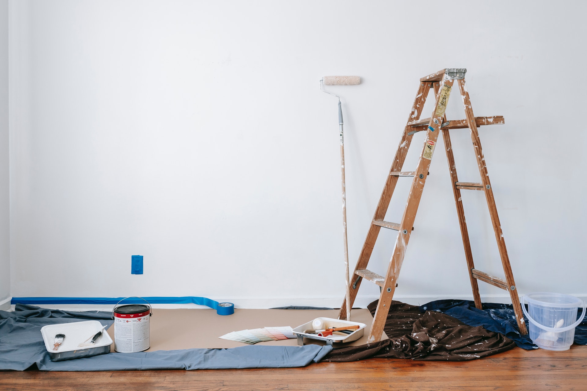 A room in the process of being painted. The wall is white. There's a ladder, a drop cloth, some paint rollers and other items.